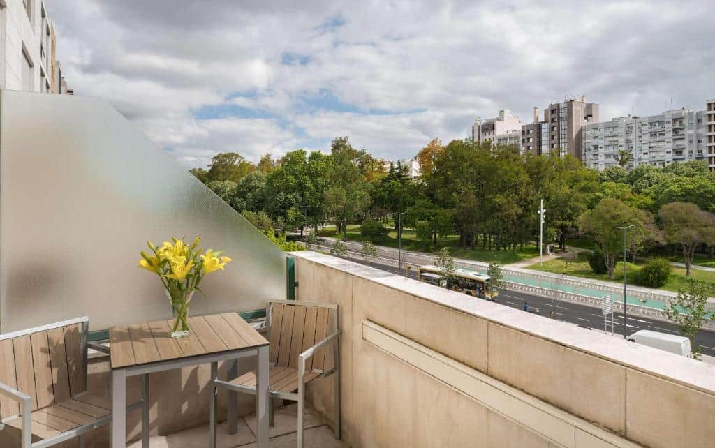 Varanda do NH Lisboa Campo Grande com vista para uma avenida arborizada, uma pequena mesa de madeira com um vaso de flores amarelas e duas cadeiras.