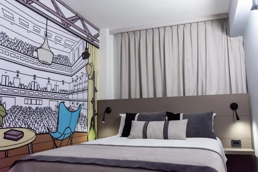 quarto do ibis Styles Buenos Aires Florida com cama de casal, mesas e candeeiros dos dois lados, janela e um papel de parede com desenhos de café em estilo cartoon