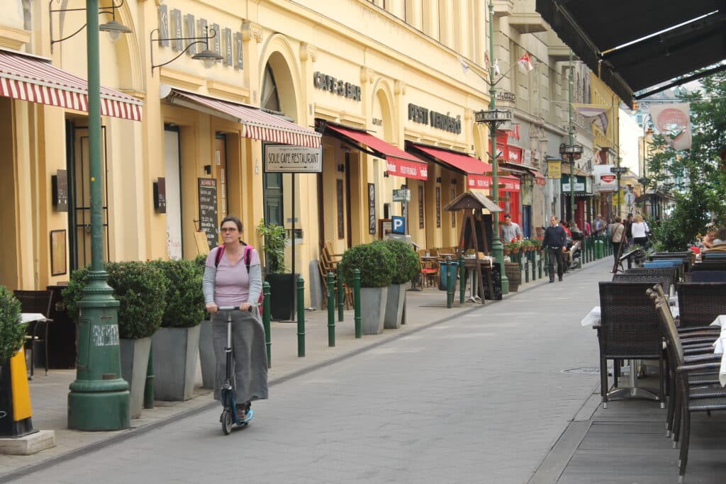 Uma mulher conduz uma trotinete elétrica numa rua pedonal de Ferencváros, repleta de cafés e restaurantes com mesas na rua.