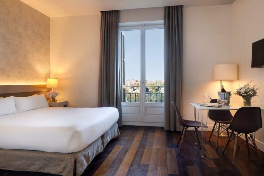Quarto do Duquesa Suites Barcelona, uma das recomendações de hotéis no Bairro Gótico em Barcelona. A cama de casal tem candeeiros nas mesas de cabeceira, uma mesa com champanhe e taças e uma porta de vidro para a varanda.
