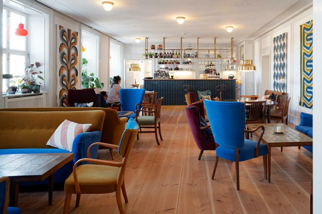 Zona comum do hotel Kanalhuset com várias mesas com cadeiras e um bar com copos e bebidas. Ao fundo, uma pessoa sentada a uma das mesas.
