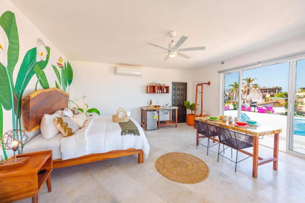 Quarto da La Casa Blanca bem iluminado com uma cama, pequena cozinha, mesa de refeições, mural na parede e portas de correr para uma varanda com vista para a piscina.