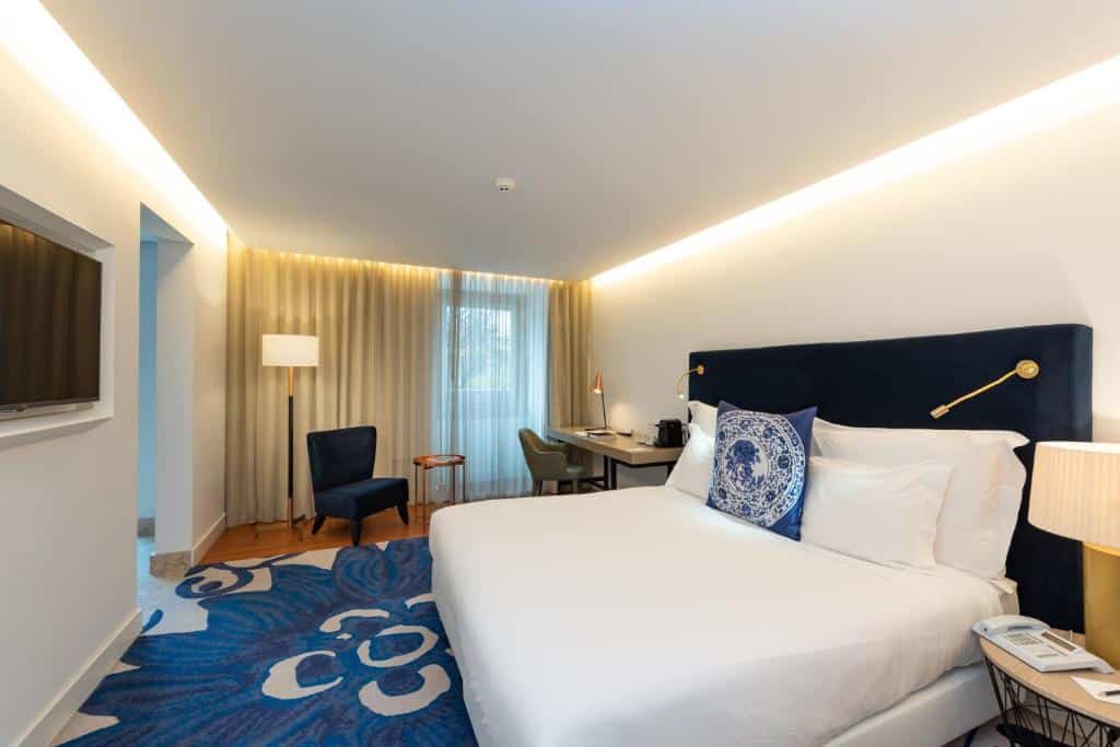 Quarto moderno no Palácio do Governador com cama grande, tapete de estampa azul, secretária, poltrona, televisão e janela do chão ao teto com cortinas.