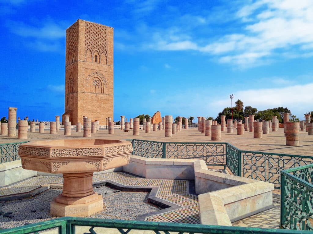 Torre Hassan com colunas de pedra e uma fonte decorativa em primeiro plano, sob um céu azul parcialmente nublado, em Rabat.