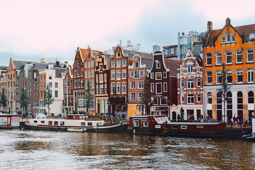 Fila de edifícios coloridos e estreitos e casas flutuantes ao longo de um canal, sob um céu nublado, num cenário de Amesterdão.