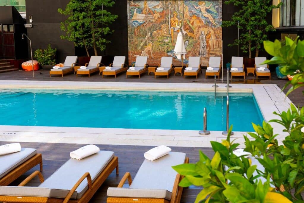 Piscina do Wellington Hotel & Spa Madrid. Espreguiçadeiras com toalhas, mesinhas e chapéus de sol estão dispostas dos dois lados, com plantas à volta. Um painel com obra de arte está no centro do local.
