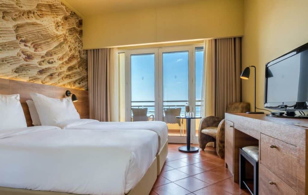 Quarto do Calheta Beach - All-inclusive - Savoy Signature com uma cama de casal do lado esquerdo da imagem em frente a cama uma cômoda com TV em cima, do lado esquerdo da cômoda uma poltrona com uma mesa redonda a frente. Representa hotéis all inclusive em Portugal.
