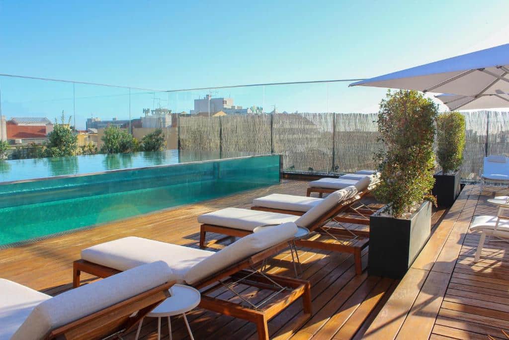 Área da piscina na cobertura com vedação de vidro, deck de madeira, espreguiçadeiras, plantas verdes e um grande guarda-sol sob um céu limpo, no Lux Lisboa Park.