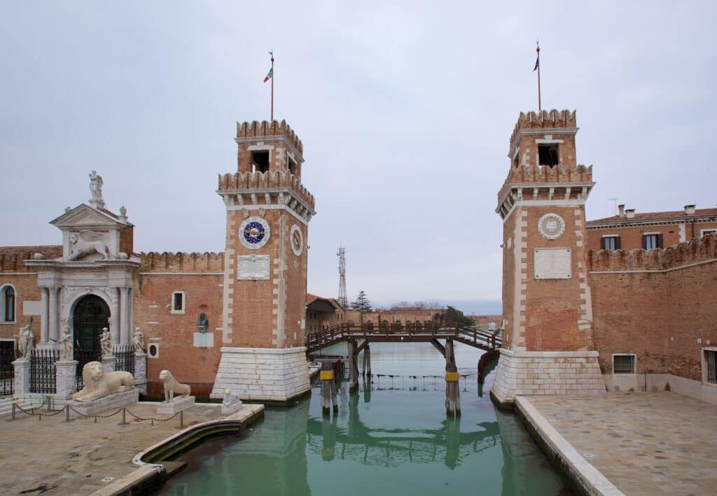 Edifício histórico de tijolo com torres e uma ponte sobre um canal numa cidade italiana