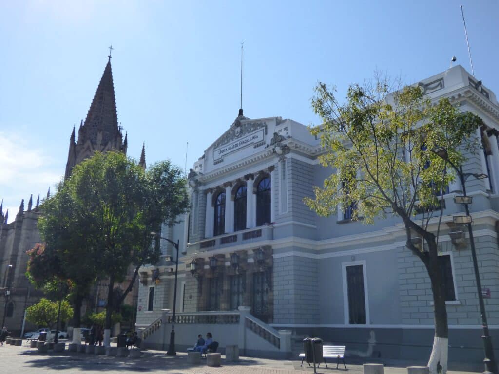Museu de Artes da Universidade de Guadalajara (MUSA), com fachada neoclássica de estilo eclético, com torres, arcadas e detalhes decorativos, em frente a uma praça com árvores e bancos.