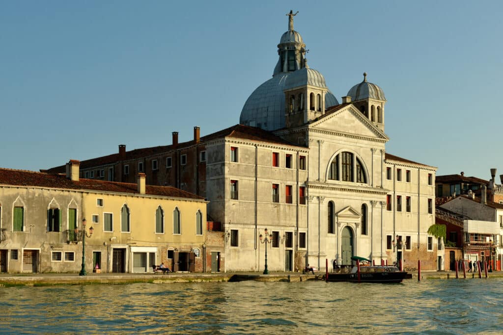 Bela igreja de cúpula dourada, com fachada imponente em estilo neoclássico, junto a um canal numa cidade italiana típica, com edifícios históricos em redor