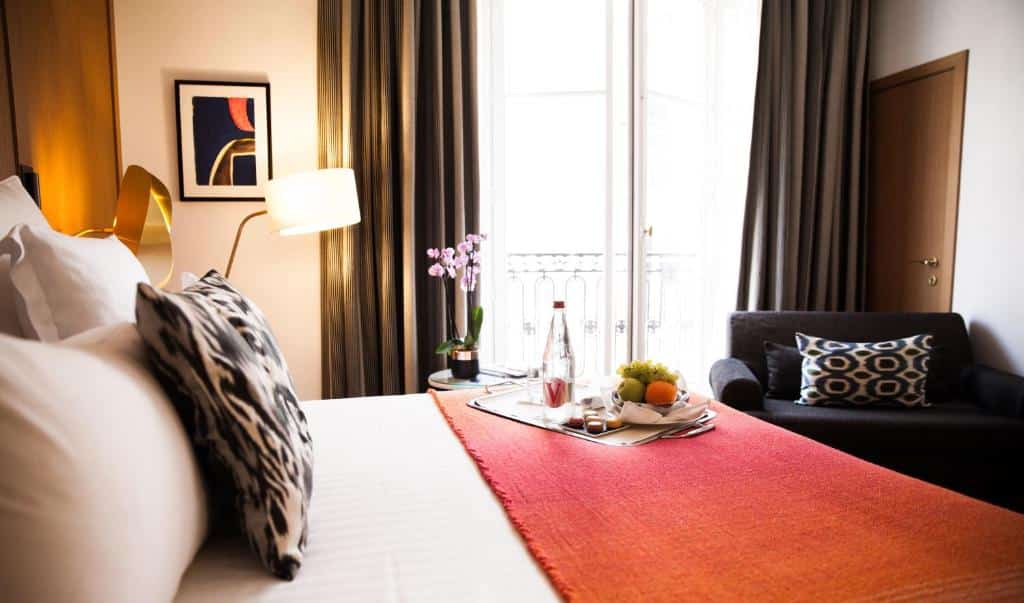 Uma cama do Hôtel Vernet Champs Elysées Paris bem feita com uma manta laranja, uma bandeja de pequeno-almoço, um pequeno sofá e uma janela com cortinas.