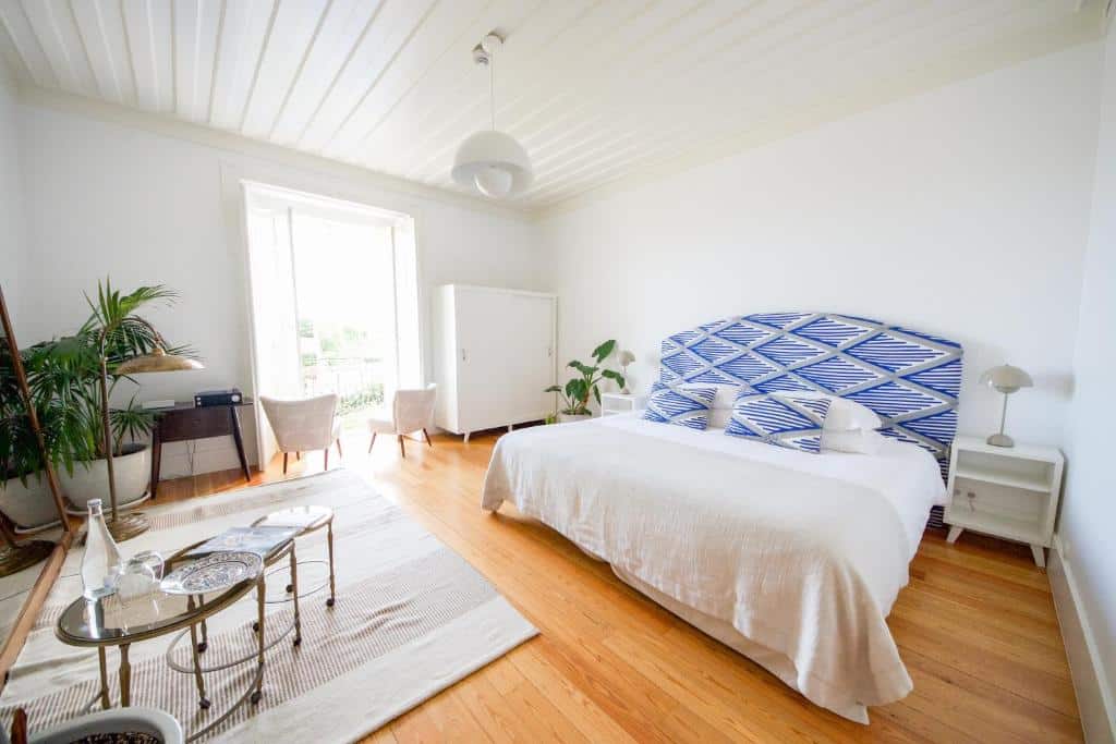 Quarto bem iluminado com pavimento de madeira, cama de casal, cabeceira estampada, mesas de apoio, cadeiras e janela grande com luz natural, na Casa Mãe em Lagos.
