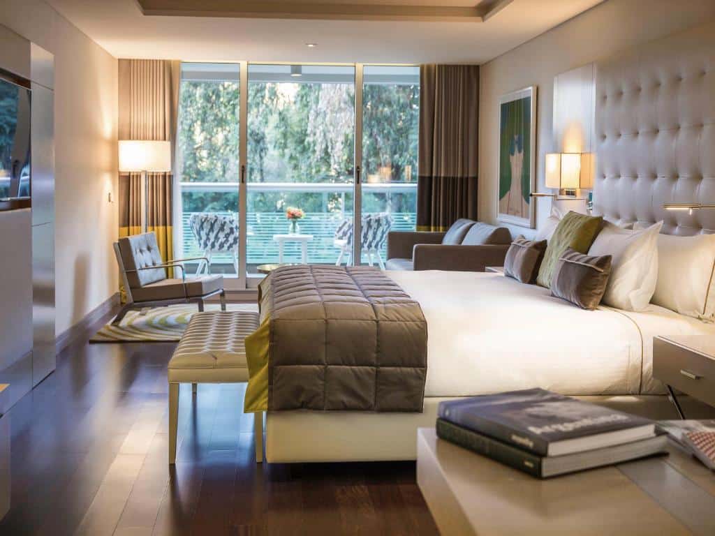 quarto do Palladio Hotel Buenos Aires MGallery com cama de casal grande, mesas de cabeceira dos dois lados e uma porta de vidro para uma varanda com mesa e cadeiras, em tons brancos e castanhos
