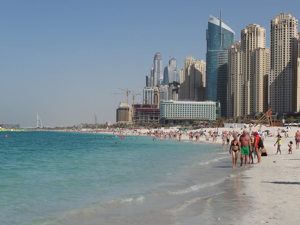 Banhistas a caminhar ao longo da Jumeirah Public Beach, com águas azul-esverdeadas e arranha-céus modernos ao fundo.