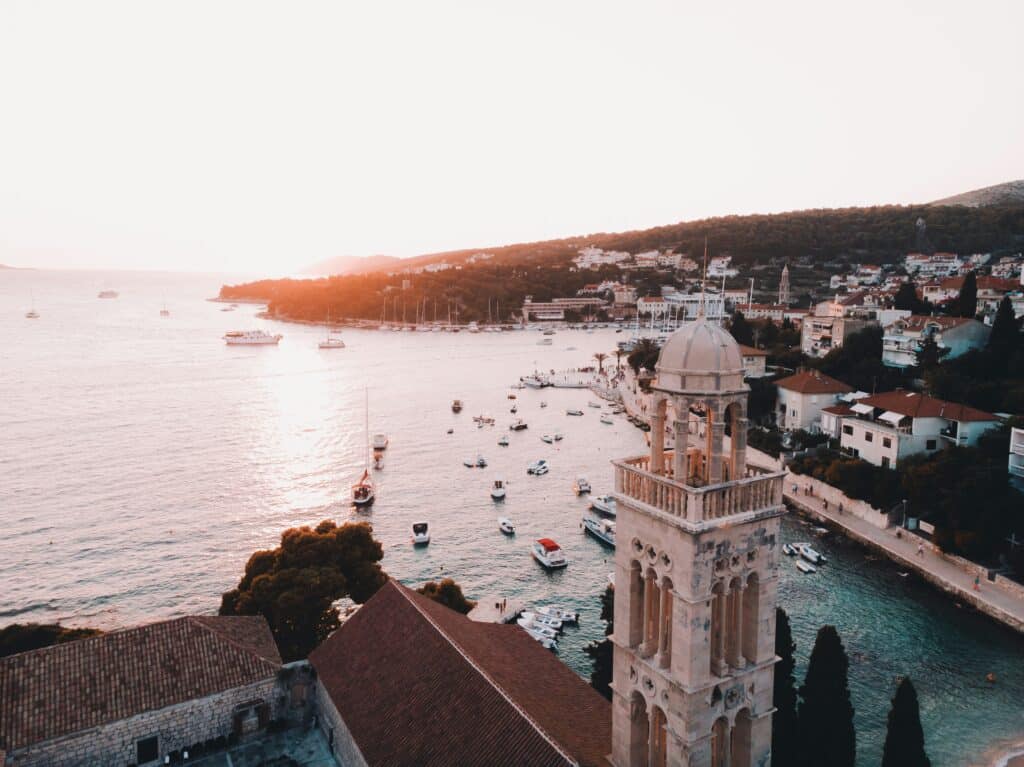 Vista aérea de Hvar, na Croácia, com uma torre de igreja proeminente, barcos na água e colinas ao fundo ao pôr do sol.