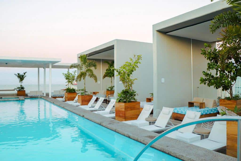 Piscina exterior do Hotel Mio Vallarta com espreguiçadeiras brancas, cabanas e vasos de plantas ao lado de um edifício moderno durante o dia.