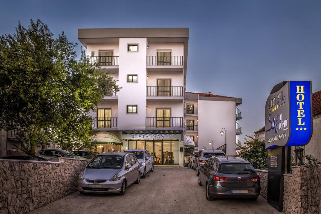 Um edifício de três andares, o Hotel Santo António de Fátima, com varandas, carros estacionados e uma entrada de restaurante ao anoitecer.