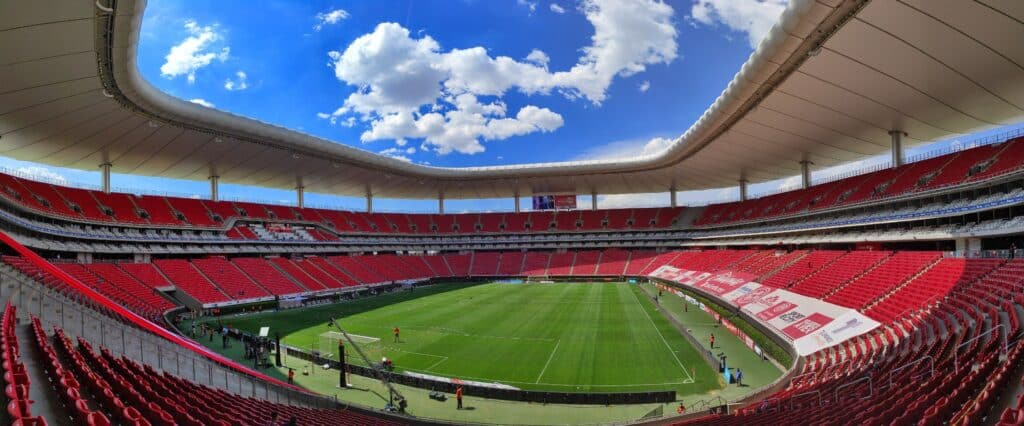 Estádio Akron, em Guadalajara, com bancadas vermelhas cheias sob céu azul com nuvens brancas.