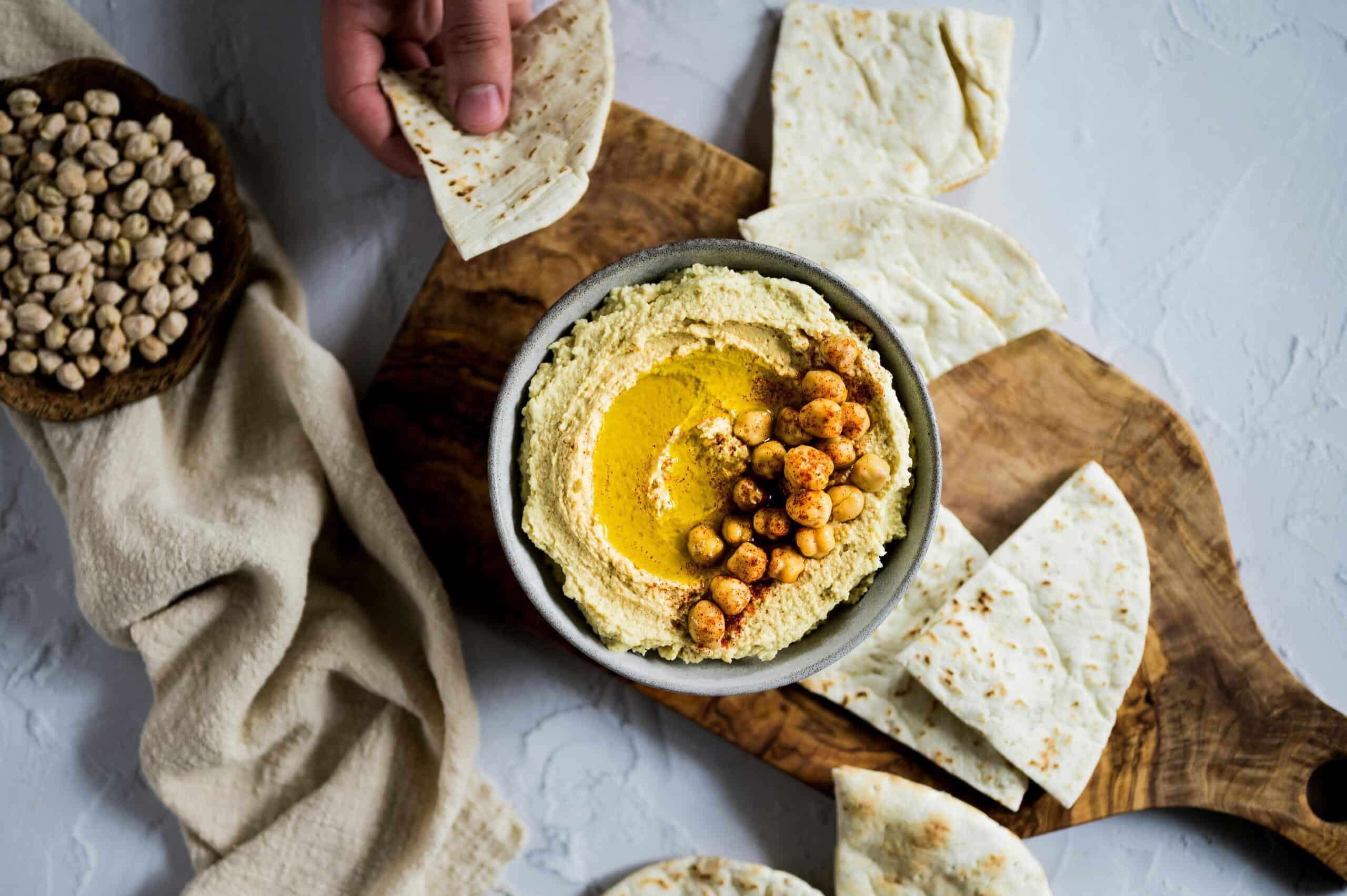 Uma tigela de húmus coberto com grão-de-bico e especiarias, sobre uma tábua de madeira com pão pita.