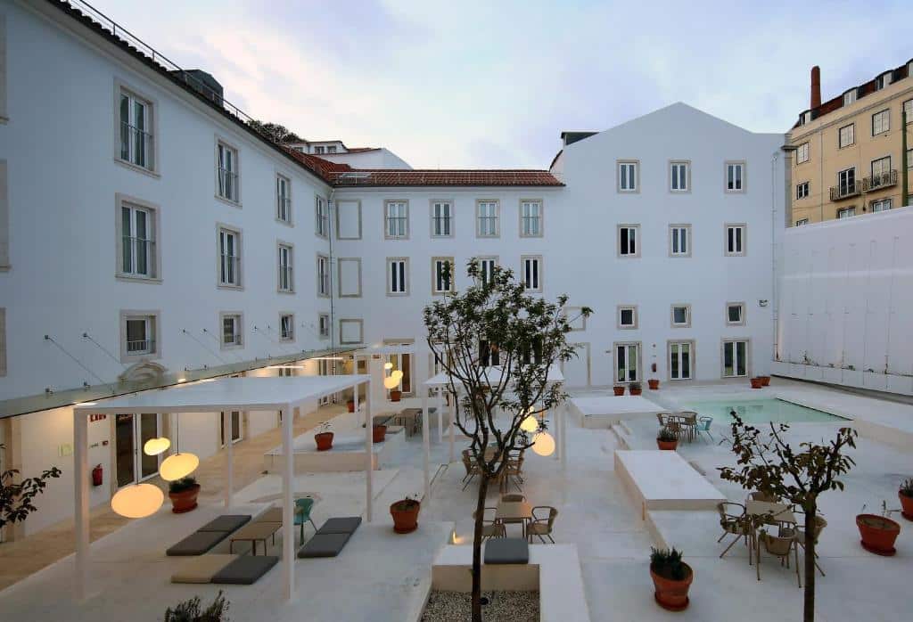 Pátio branco e moderno do Hotel Convento do Salvador, com zonas de estar, vasos de plantas, candeeiros suspensos e uma piscina retangular, rodeado de edifícios