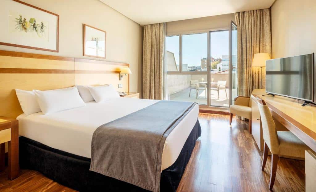 Quarto do Ilunion Alcalá Norte, um dos melhores hotéis em Madrid. Uma cama de casal tem mesinhas de cabeceira e candeeiros dos dois lados e fica em frente a uma secretária com cadeira e TV. Ao fundo, uma poltrona e um candeeiro de pé ficam junto à porta de vidro que ocupa uma parede e dá acesso à varanda com mesinha e cadeiras.
