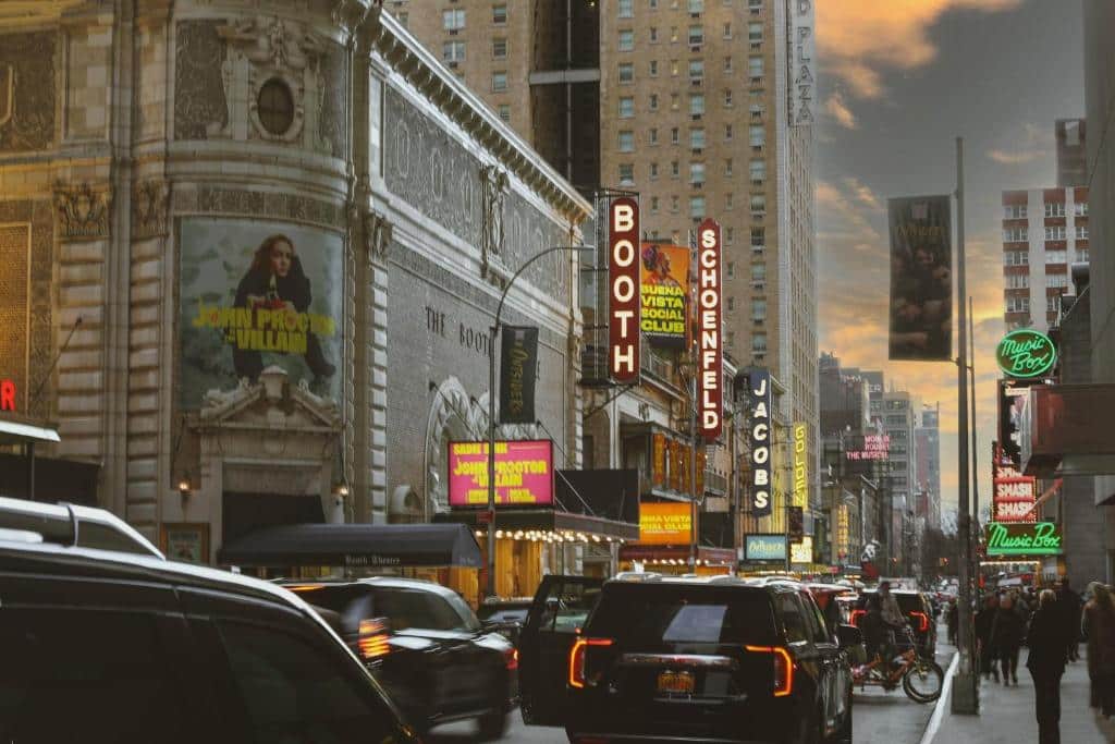 Rua da Broadway ao pôr do sol, com carros, pessoas e letreiros iluminados de teatros, entre eles o Booth e o Schoenfeld.