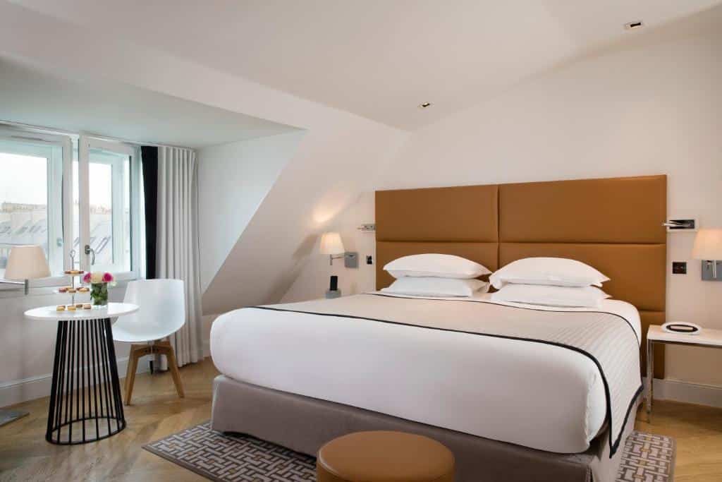 Quarto do Hôtel R de Paris com cama grande, dois candeeiros de cabeceira e uma mesa redonda junto a uma janela com cortinas.