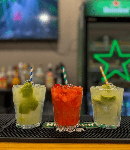 Três bebidas com palhinhas às riscas no Maria Bonita, sobre um balcão de bar, com garrafas ao fundo.