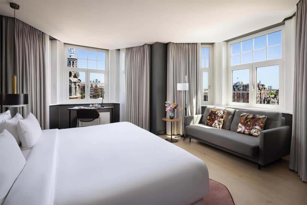 Quarto do NH Collection Amsterdam, que ilustra onde ficar em Amesterdão: cama grande, sofá cinzento, secretária e janelas com vista para uma paisagem urbana com uma torre de relógio.