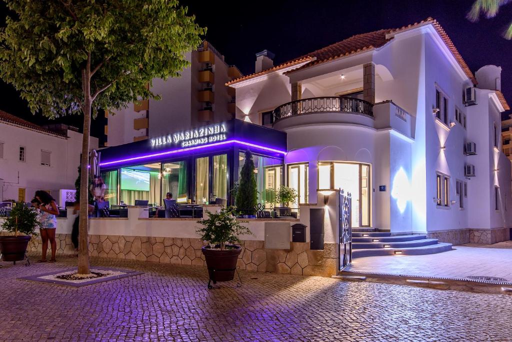 Edifício branco moderno, propriedade da Villa Mariazinha Charming Hotel, com letreiro luminoso ao anoitecer, lugares ao ar livre e árvores ao longo de um passeio em pedra.