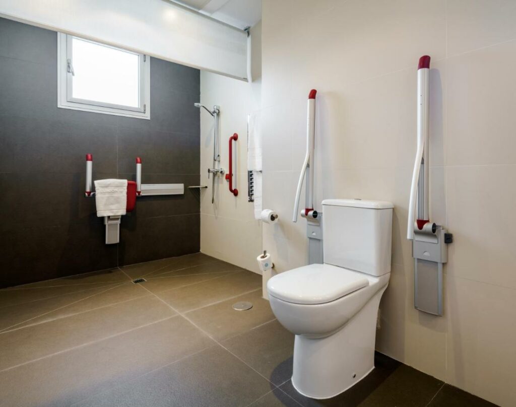Casa de banho adaptada para pessoas com mobilidade reduzida no Ilunion Alcalá Norte. A sanita tem barras de apoio dos dois lados e rolos de papel higiénico entre a zona de banho e a sanita. Ao lado há barras de apoio à volta do chuveiro e uma toalha branca pendurada no assento de banho. Há uma pequena janela na parede por cima.