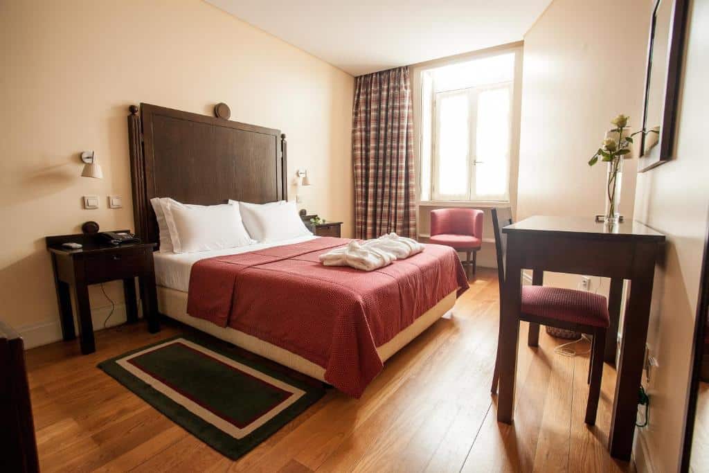 Quarto do Hotel Bracara Augusta com cama de casal, colcha vermelha, pavimento em madeira, secretária, cadeira e mesas de cabeceira.