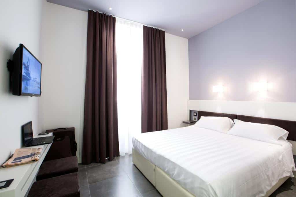 quarto amplo do Crosti Hotel, um dos hotéis baratos em Roma, com cama enorme, mesa de cabeceira e candeeiro de ambos os lados, janela grande até ao teto com cortinados escuros, uma mesa com pufes e televisão em frente