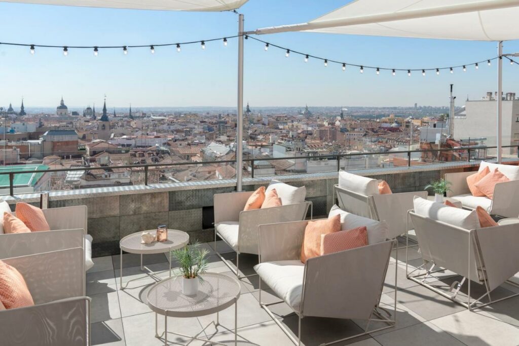 Terraço do Vincci Capitol com vista para a cidade. Diversas poltronas brancas com almofadas cor de rosa estão dispostas com mesinhas entre elas. Há luzes penduradas nas tendas brancas por cima e um muro com proteção de vidro fica na beira do local.