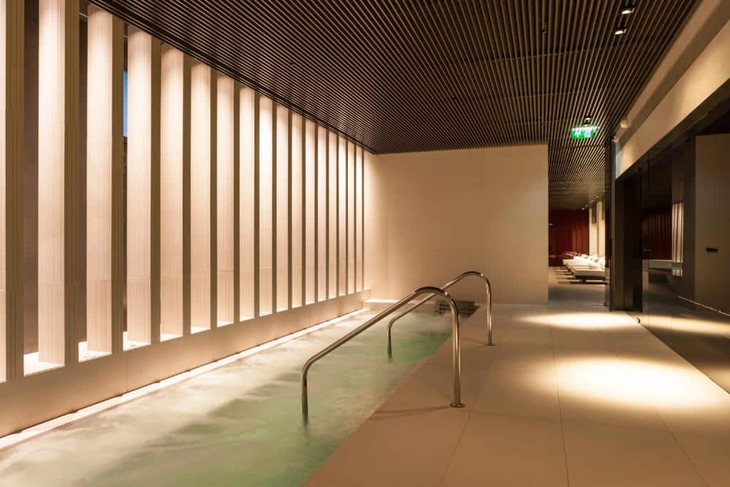 Piscina de spa interior do Kozmo Hotel Suites &amp; Spa com corrimões de metal, janelas verticais altas e iluminação moderna ao longo de um corredor elegante.