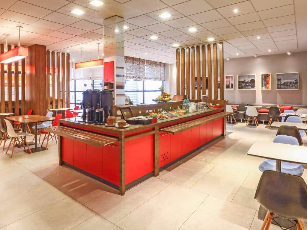 área do pequeno-almoço do ibis Buenos Aires Obelisco, moderna e bem iluminada, com uma ilha de buffet em vermelho e mesas e cadeiras coloridas