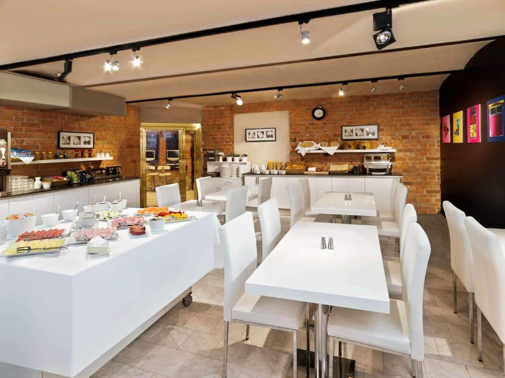 Sala de pequeno-almoço do Ibis Styles Stockholm Odenplan com mesas e cadeiras e buffet com fruta e aperitivos.