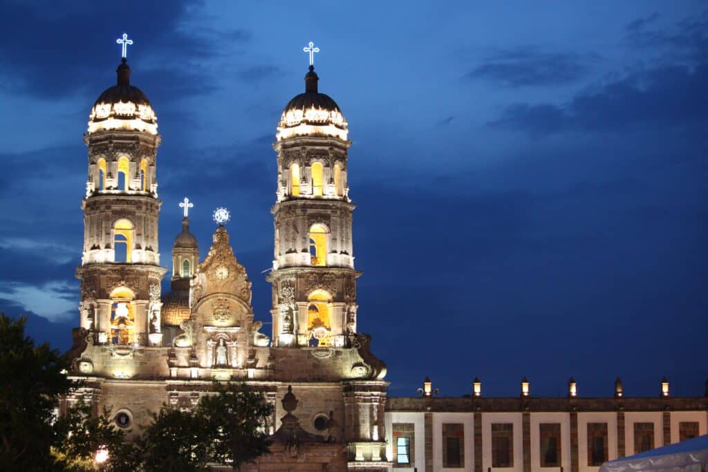 Templos com torres iluminadas no centro histórico da cidade à noite; trata-se da Basílica de Nossa Senhora de Zapopan.