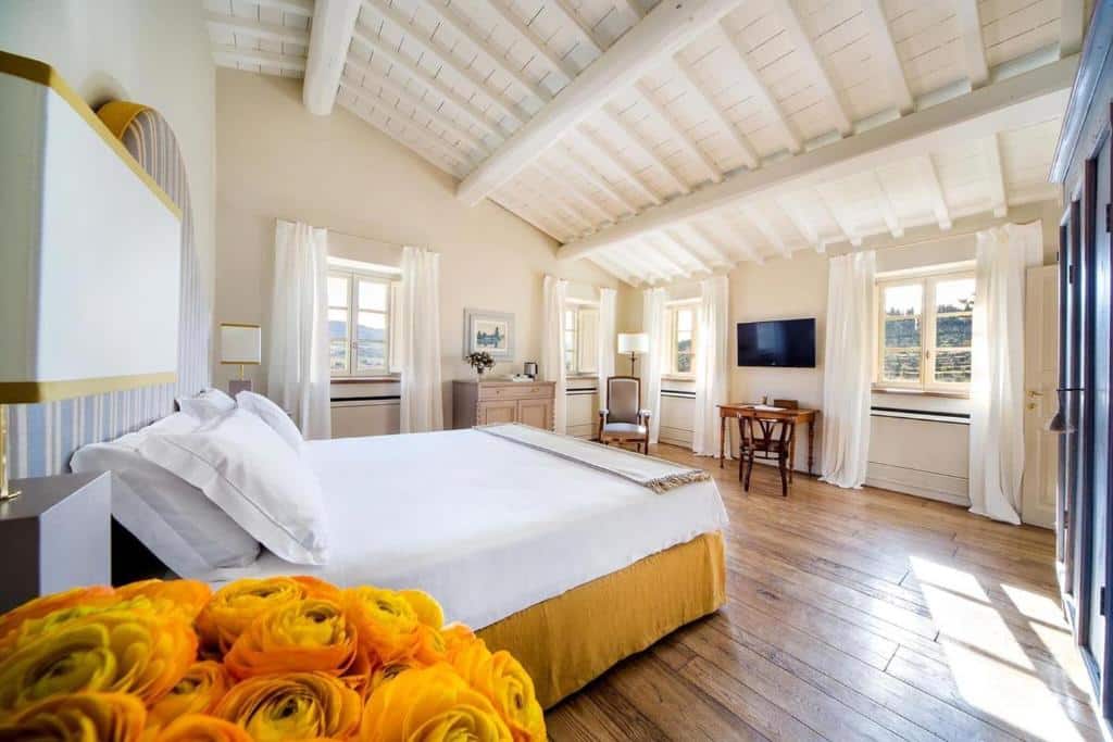 Quarto duplo deluxe do Borgo Del Cabreo, com 30 m², cama de casal grande, flor amarela, TV, mesas retro, poltrona e uma cadeira, com bastante espaço entre a cama e as paredes