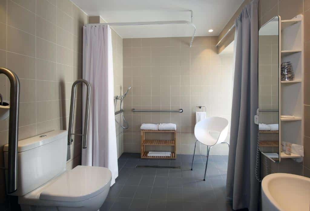 Casa de banho adaptada do Hotel Convento do Salvador com barras de apoio junto à sanita e duche sem resguardo de vidro, com banco para sentar.