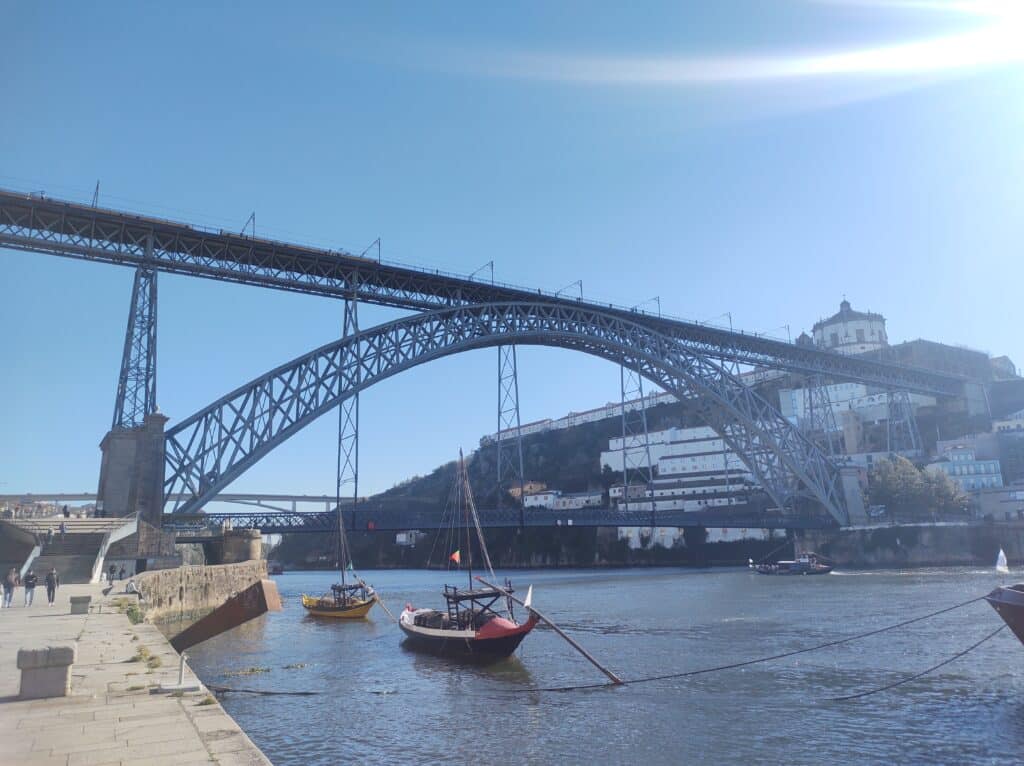 Ponte D. Luís I no Porto durante o dia, com barcos no rio e a ponte ao fundo. Representa o que fazer à noite no Porto.