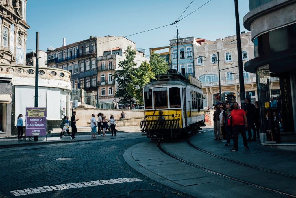 Um elétrico amarelo e branco circula por uma esquina no Porto, com vários peões por perto num dia claro e soalheiro. Representa o que fazer à noite no Porto.