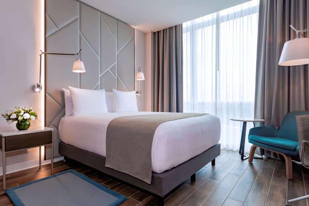 Quarto no NH Cancun Airport com cama de casal, varanda, poltrona, candeeiros suspensos, vasos de flores e chão de madeira