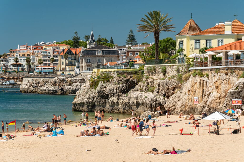 Pessoas a apanhar sol e a nadar na Praia da Rainha, em Cascais, com falésias rochosas e edifícios ao fundo, sob um céu azul.