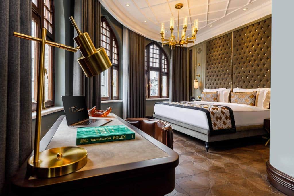 Quarto do Cronton Design Hotel com uma cama grande, cabeceira capitonê, secretária com livros, candeeiro dourado e janelas em arco com cortinas.