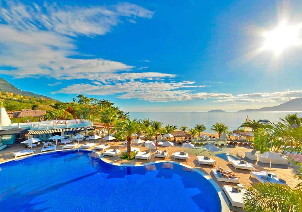 Piscina de um hotel com vista para o mar azul, espreguiçadeiras brancas e palmeiras tropicais em Ilhabela