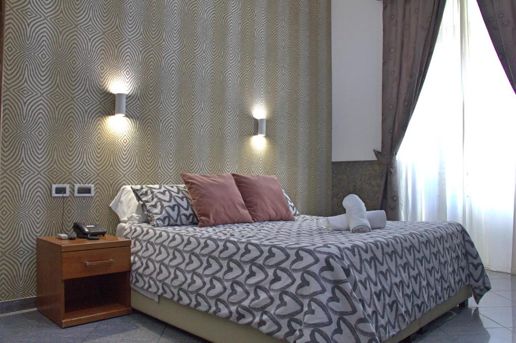 quarto do Hotel Ciao, um dos hotéis baratos em Roma, com cama de casal com colcha em forma de coração, almofadas em tons de vermelho-pálido a condizer com os cortinados da janela grande com vista para a rua, mesas de cabeceira e candeeiros de ambos os lados