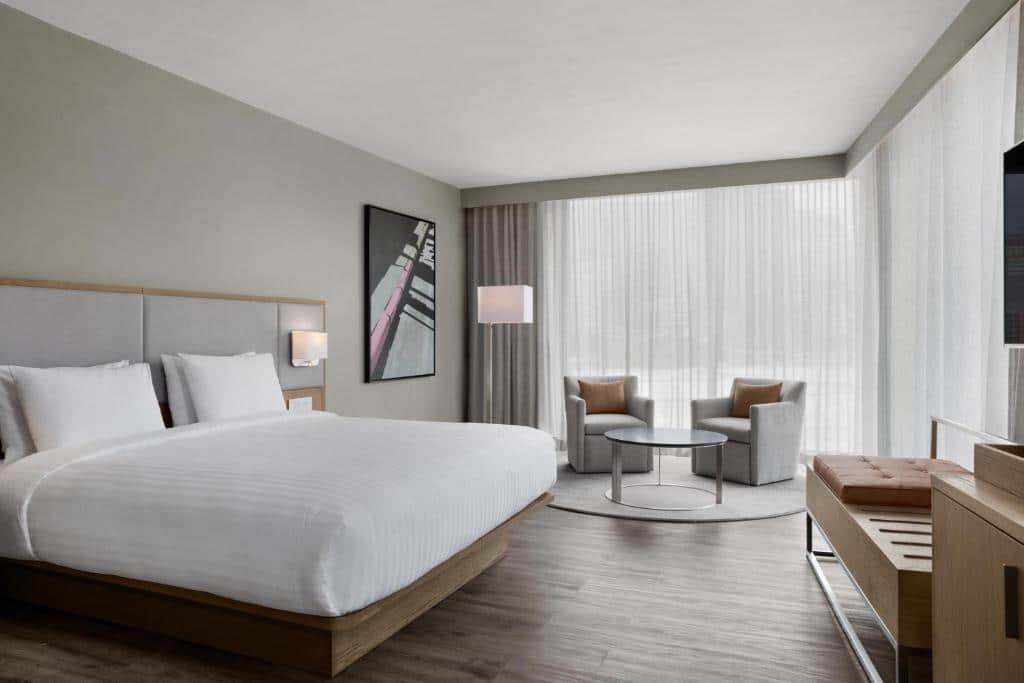 Quarto do AC Hotel by Marriott com cama grande, duas cadeiras junto a uma janela, mesa pequena, obras de arte e decoração em cores neutras.