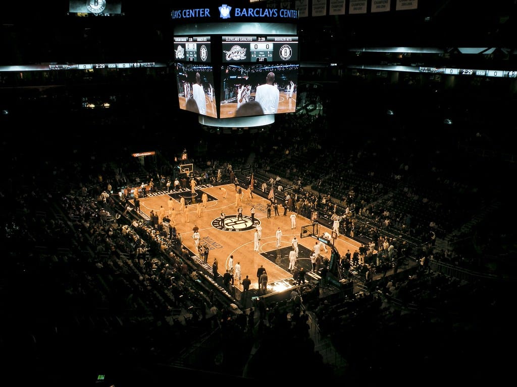 Arena coberta com um campo de basquetebol ao centro, bancadas com espectadores e um grande ecrã suspenso.